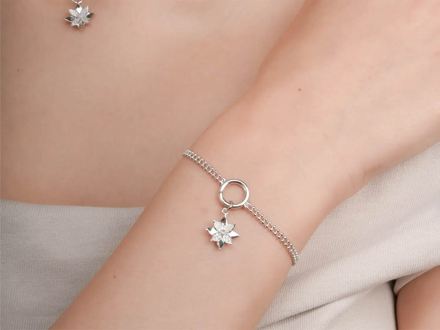 Close-up of a person’s wrist wearing a silver beaded chain bracelet with a circular clasp and a silver poinsettia flower charm. A matching silver poinsettia pendant necklace is partially visible in the background.
