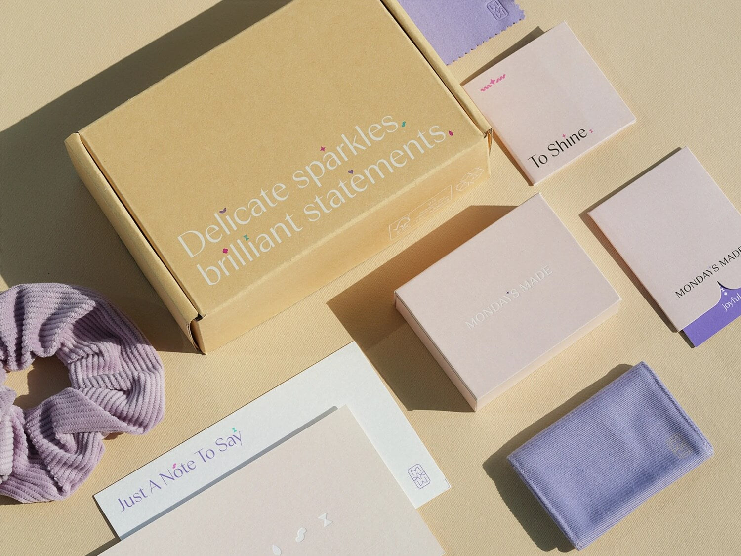 Mondays Made Shipping box with text "Delicate sparkles, brilliant statements", polishing cloth, jewelry box, purple jewelry pouch, purple scrunchie and message card.
