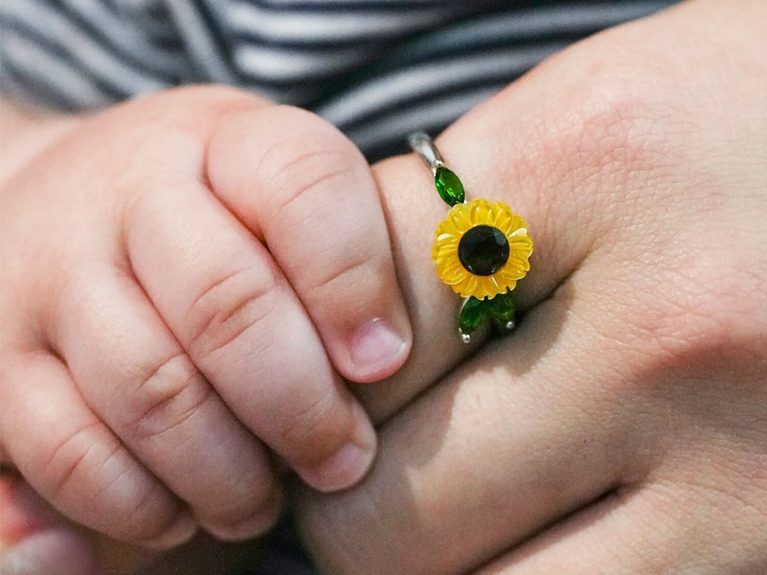 Close-up of a child's hand holding onto an adult hand wearing our Carved Sunflower Ring.