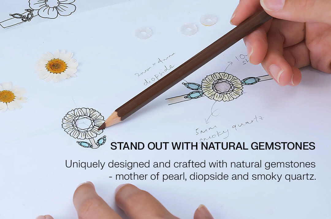 Hand holding a pencil with gemstone accents on a drawing of flowers, text about natural gemstones. "Stand out with natural gemstones, uniquely designed and crafted with natural gemstones - mother of pearl, diopside and smoky quartz."