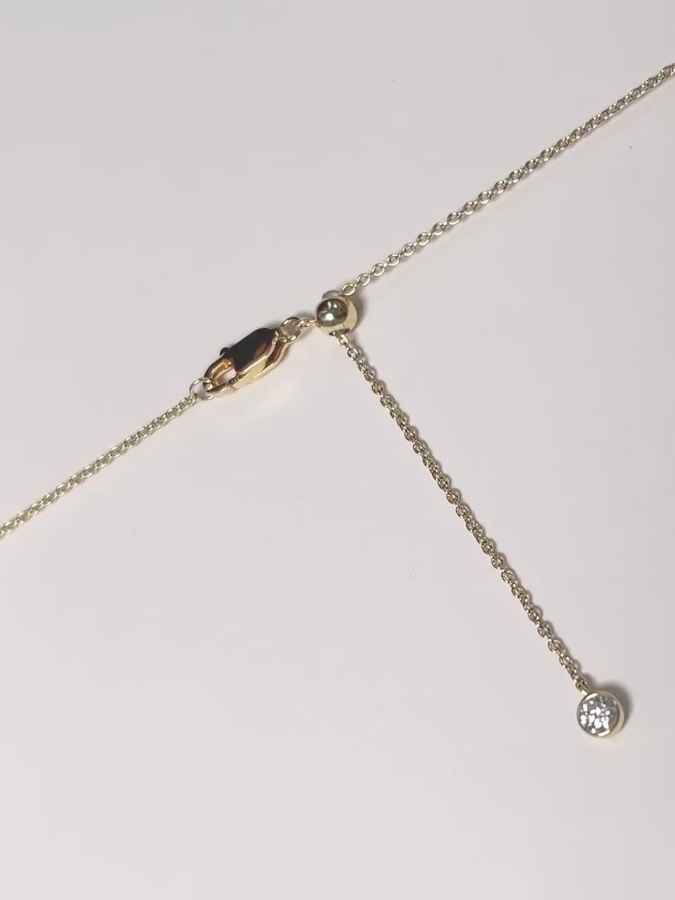 A close-up video demonstrating how to adjust the length of a gold necklace. The camera focuses on the clasp area, which features a lobster clasp, a small gold slider ball, and a delicate chain end with a small charm. The video then shows the end of the chain being gently pulled through the slider ball, illustrating the smooth adjustment mechanism.