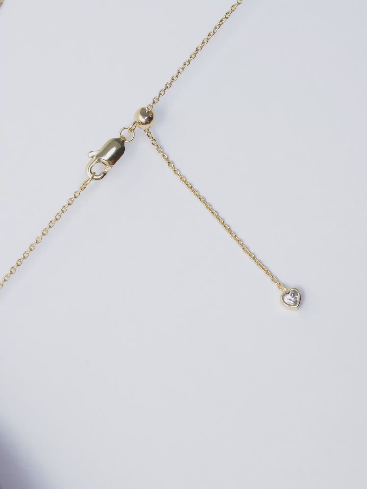 A close-up video demonstrating how to adjust the length of a gold necklace. The camera focuses on the clasp area, which features a lobster clasp, a small gold slider ball, and a delicate chain end with a small, heart-shaped charm. On-screen text instructs: "Hold onto the slider ball" and "Pull the chain to adjust necklace length." The video then shows the end of the chain being gently pulled through the slider ball, illustrating the smooth adjustment mechanism.