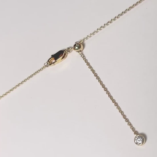 A close-up video demonstrating how to adjust the length of a gold necklace. The camera focuses on the clasp area, which features a lobster clasp, a small gold slider ball, and a delicate chain end with a small charm. The video then shows the end of the chain being gently pulled through the slider ball, illustrating the smooth adjustment mechanism.