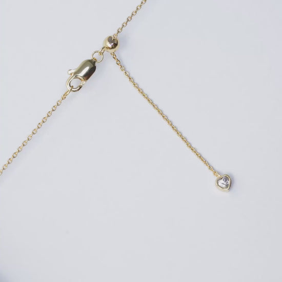 A close-up video demonstrating how to adjust the length of a gold necklace. The camera focuses on the clasp area, which features a lobster clasp, a small gold slider ball, and a delicate chain end with a small, heart-shaped charm. On-screen text instructs: "Hold onto the slider ball" and "Pull the chain to adjust necklace length." The video then shows the end of the chain being gently pulled through the slider ball, illustrating the smooth adjustment mechanism.